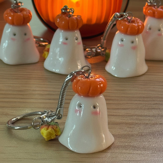 A set of cute ceramic bell keychains designed as ghosts with orange pumpkins on their heads, displayed on a wooden table with a glowing orange pumpkin lantern in the background.
